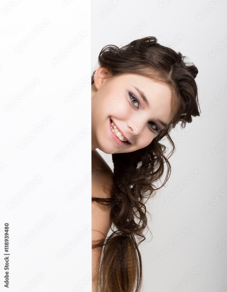 Smiling happy young woman standing behind and leaning on a white blank billboard or placard, expresses different