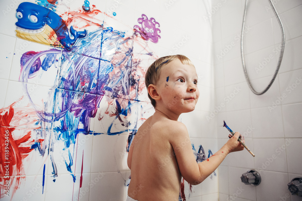 Fototapeta premium Portrait of cute adorable white Caucasian little boy playing and painting with paints on wall in bathroom having fun, lifestyle childhood concept
