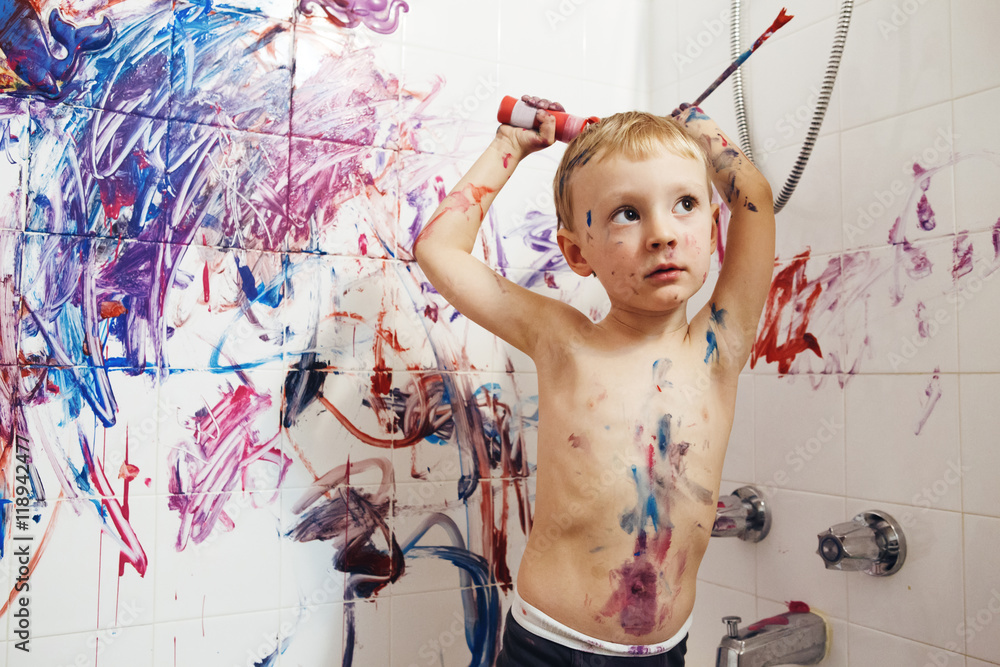 Fototapeta premium Portrait of cute adorable white Caucasian little boy playing and painting with paints on wall in bathroom having fun, lifestyle childhood concept