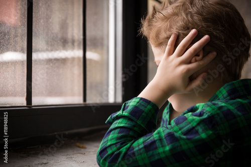 Little boy standing behind the window in sad mood. Sad Teenager looking in the Window and closing his ears with hands. Unhappy child in a plaid shirt. Alone at home. Upset.