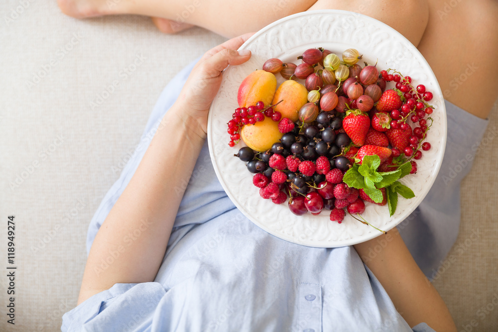 Top view on woman sitting cozy on the couch and enjoying delicious summer fruit and berries. Girl holding white plate with apricots, raspberries, strawberries, blackcurrant, blueberries on her knees Top view on woman sitting cozy on the couch and enjoying delicious summer fruit and berries. Girl holding white plate with apricots, raspberries, strawberries, blackcurrant, blueberries on her knees