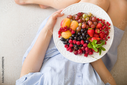Top view on woman sitting cozy on the couch and enjoying delicious summer fruit and berries. Girl holding white plate with apricots, raspberries, strawberries, blackcurrant, blueberries on her knees