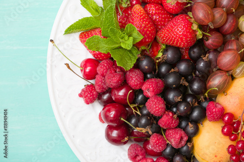 Assortment of juicy fruits on white plate and turquoise table background. Strawberries  blueberries  raspberries  gooseberry  blackcurrant decorated with mint for summer dessert or snack.