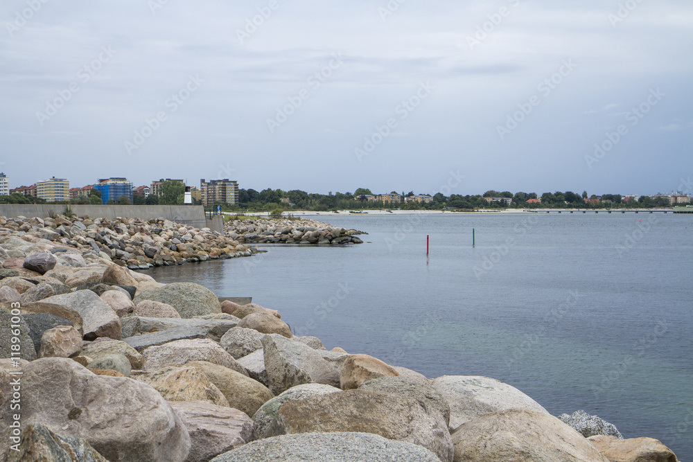 Stenig strandkant vid hamninlopp med utsikt över stad