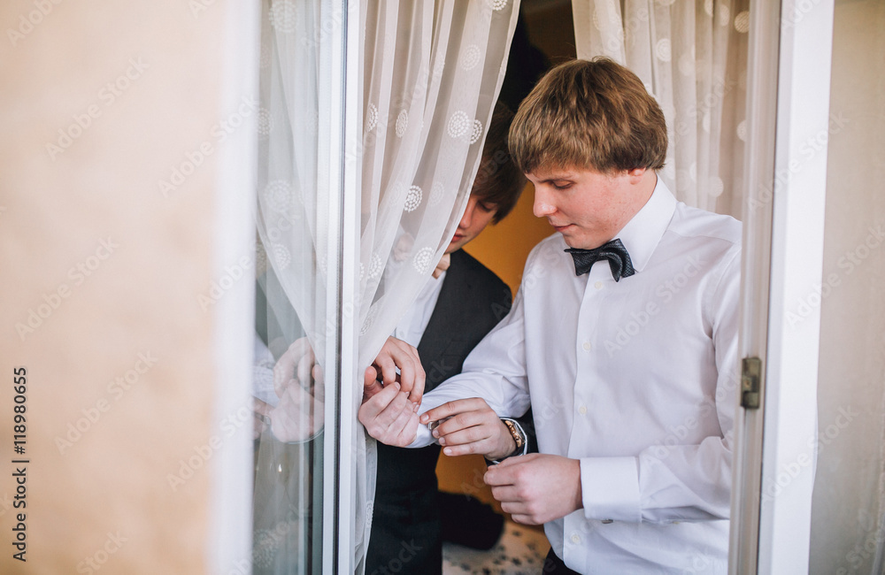 Fototapeta premium Groom and his friends prepare for the wedding. Grooms man helps the groom to put on black cufflinks on white sleeve of the shirt. Details of the morning of the wedding day