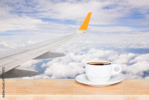 Wooden table and cup coffee with blurred airport as seen through window of an aircraft after takeoff. Wing of the plane on airport in background photo