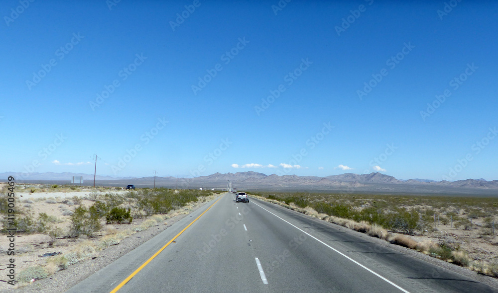 Autobahn in der Mojave-Wüste/Lange, gerade Autobahn durch die Mojave-Wüste in Kalifornien, zweispurige Fahrbahn mit Markierung und Standstreifen, bergige Landschaft und blauer Himmel