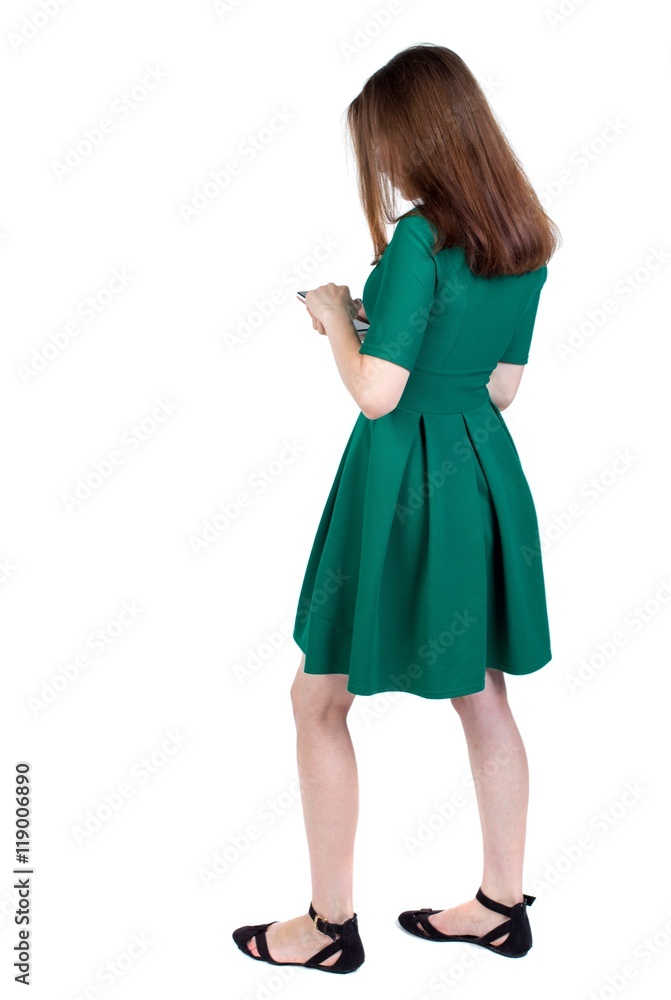 back view of standing young beautiful woman using a mobile phone or tablet computer. slender brunette in a green short dress stands sideways looking at the phone. back view of standing young beautiful woman using a mobile phone or tablet computer. slender brunette in a green short dress stands sideways looking at the phone.