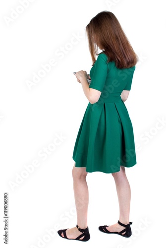 back view of standing young beautiful woman using a mobile phone or tablet computer. slender brunette in a green short dress stands sideways looking at the phone.