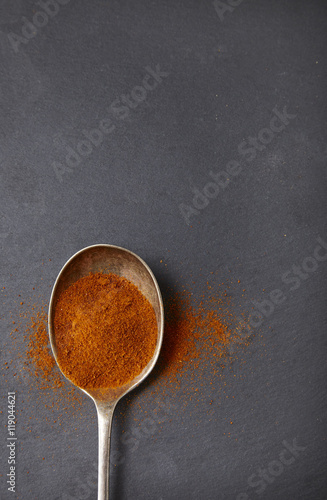 An old metal spoon full of chili powder spices on a rustic slate background forming a page border