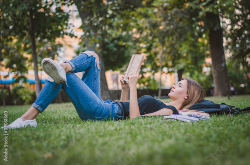 girl student in a park lying on the grass and reading a book, lying next to her backpack and a stack of notebooks. Education concept