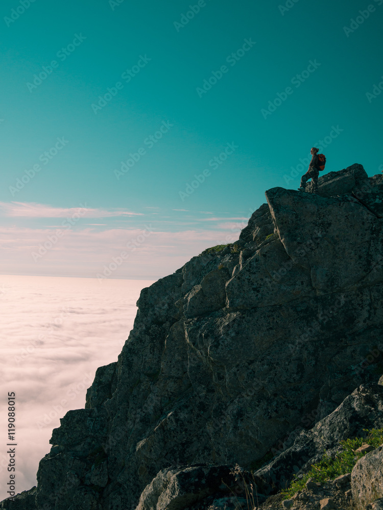 Toned image adult woman with a backpack stands on the edge of a cliff and looking at the sunrise against the blue sky and thick clouds floating down