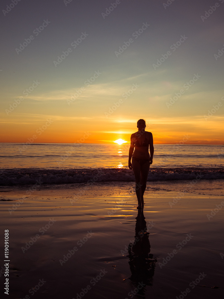 Toned image of a silhouette of an adult woman walking on the beach at sunset and colored background of the waves