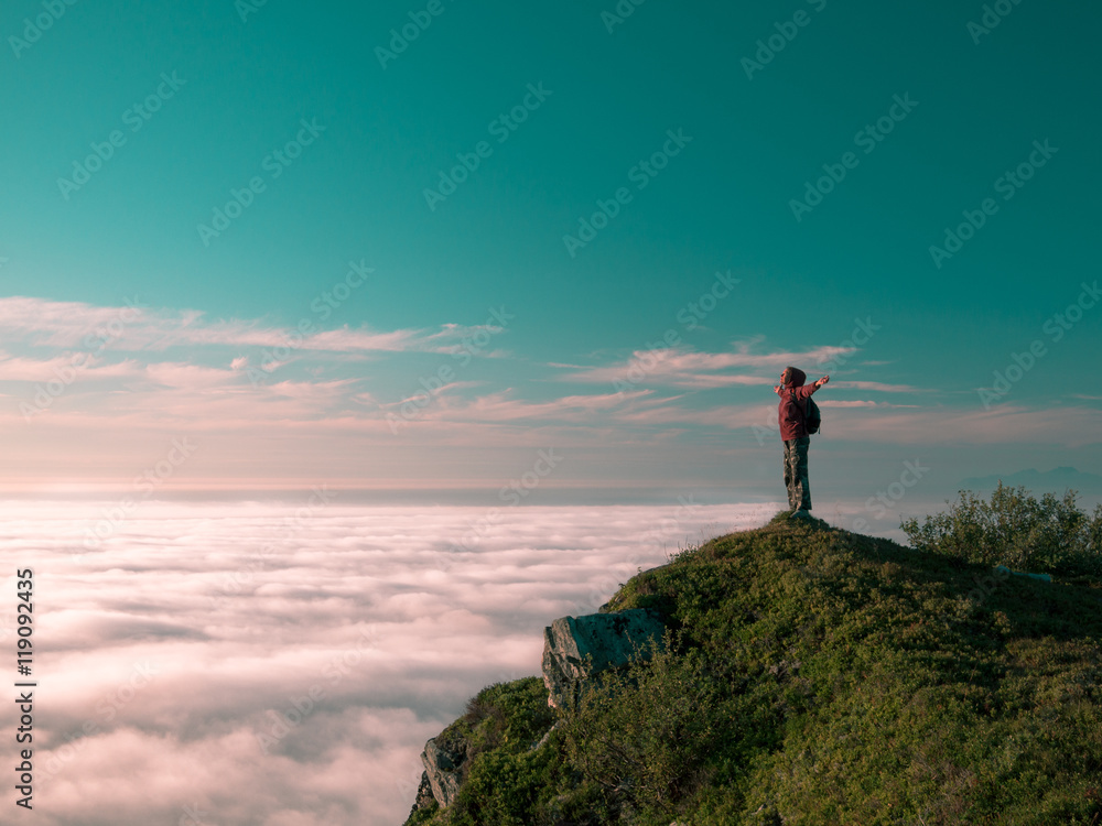 Toned image adult woman with a backpack stands on the edge of a cliff and looking at the sunrise against the blue sky and thick clouds floating down