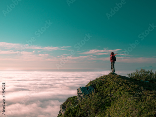 Toned image adult woman with a backpack stands on the edge of a cliff and looking at the sunrise against the blue sky and thick clouds floating down