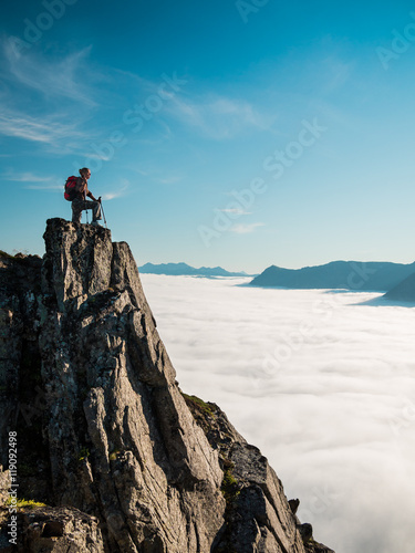 Adult woman with a backpack and Alpenstocks stands on the edge of a cliff and looking at the sunrise against the blue sky and thick clouds floating down