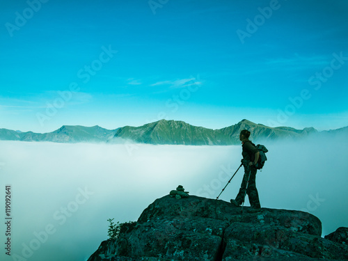 Toned image of an adult woman standing on top of a mountain with a backpack and Alpenstocks against mountains in a fog and the blue sky