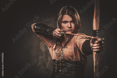 Murais de parede Serious viking woman with bow and arrow in a traditional warrior clothes, posing on a dark background