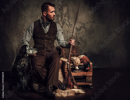 Obraz na plátně Handsome hunter with a english setter and shotgun in a traditional shooting clothing, sitting on a dark background