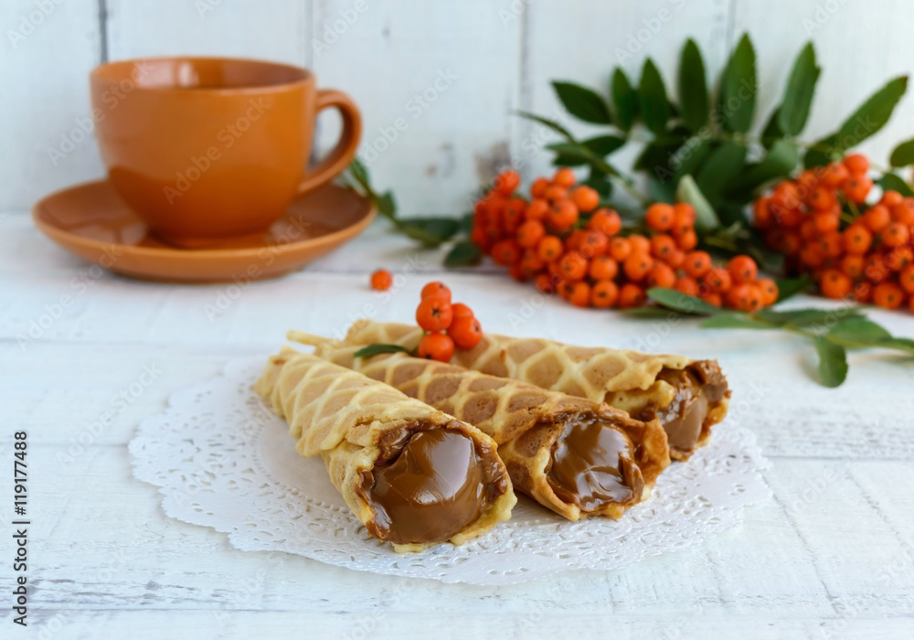 Homemade honey wafers rolled into a cone, filled with caramel (condensed milk), a cup of tea, rowan berries on a white background. Close up