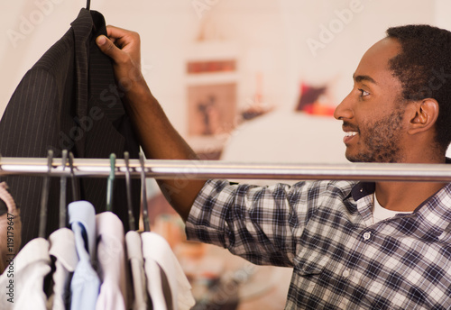 Handsome young man standing inside wardrobe going through rack of different clothes hanging, fashion concept