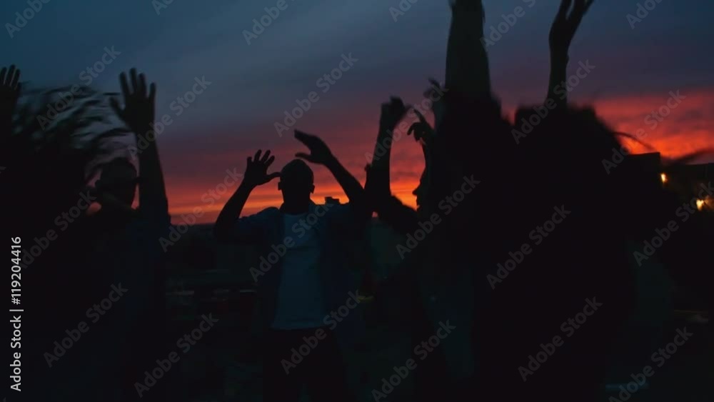 Silhouettes of group of young people dancing and raising their arms up in the air against beautiful sunset sky at rooftop party Silhouettes of group of young people dancing and raising their arms up in the air against beautiful sunset sky at rooftop party