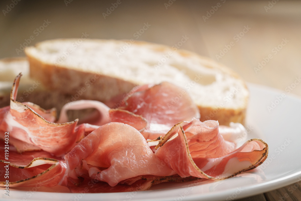 speck slices on white plate with ciabatta on old wood table speck slices on white plate with ciabatta on old wood table