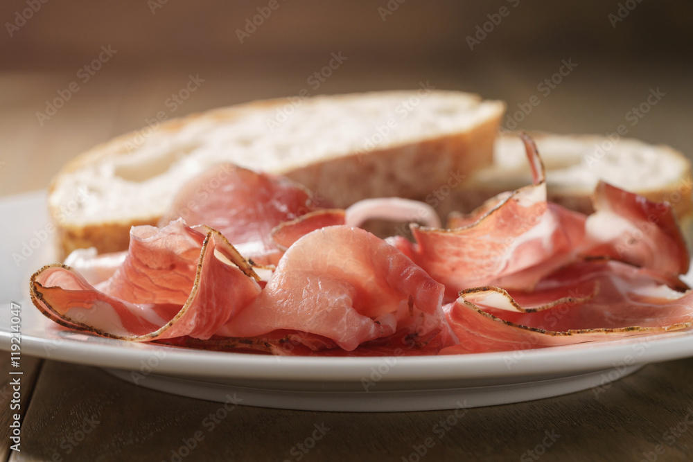 speck slices on white plate with ciabatta on old wood table