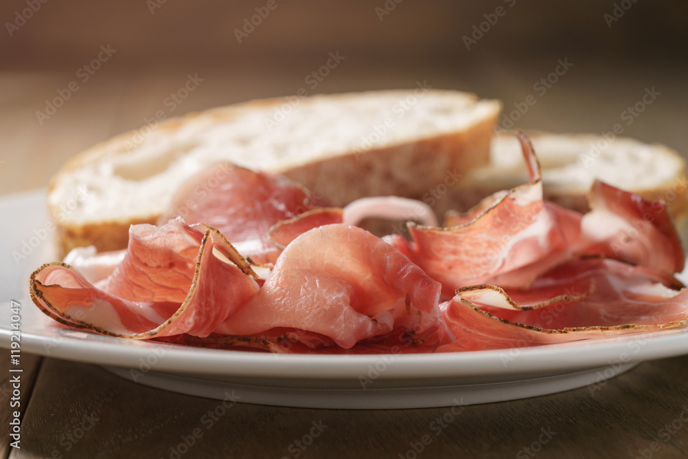 speck slices on white plate with ciabatta on old wood table speck slices on white plate with ciabatta on old wood table