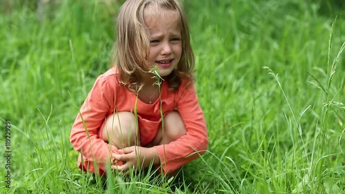 Little girl in red dress crying, because she got burnt with nettle. True sincere weeping and emotions of little girl. Little girl sits on grass and crying. Real situation, pain caused by chemical burn