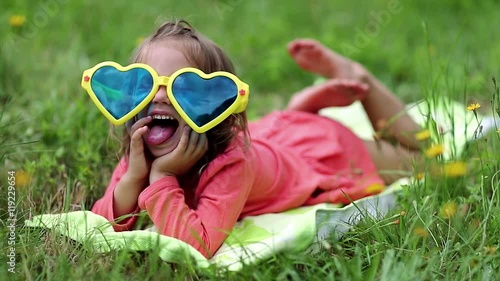 Beautiful little girl in big sunglasses lies on the grass. Funny little girl in glasses lies on the green grass and looks at the camera photo