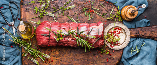 Raw roast beef  with herbs tied with a rope with cooking ingredients, oil  and spices on rustic background, top view, banner