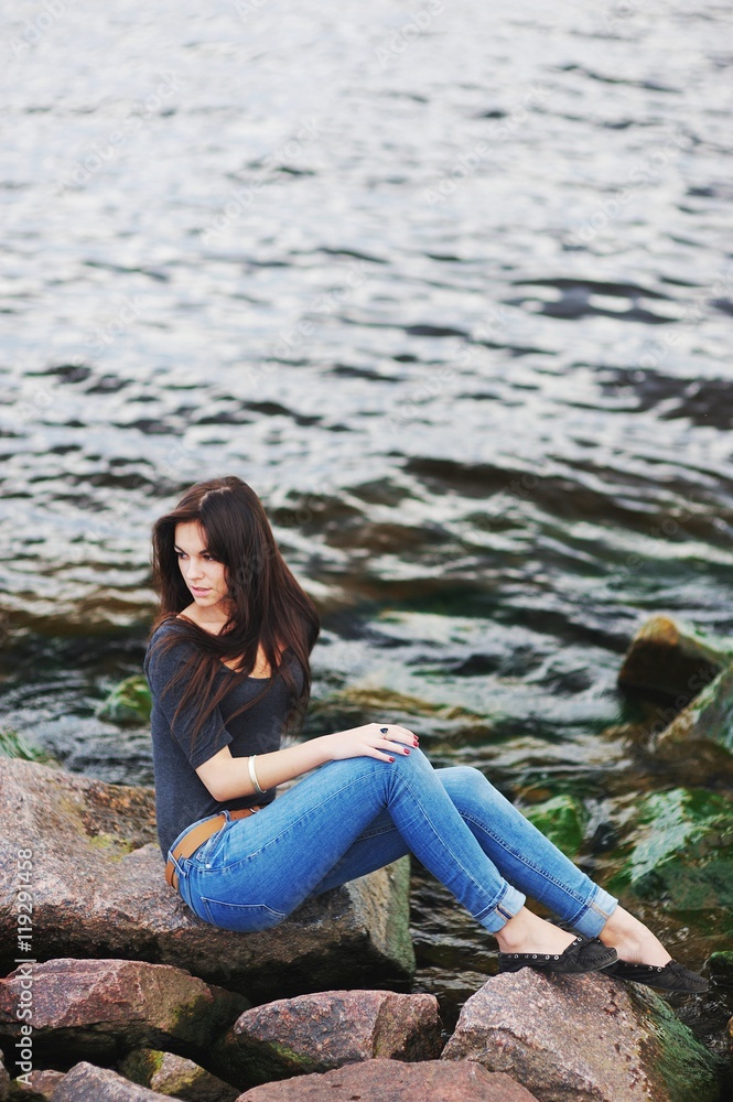 Cute long-haired girl in blue jeans and a black blouse poses on the rocks by the sea, the emerald green waves lapping against the shore.