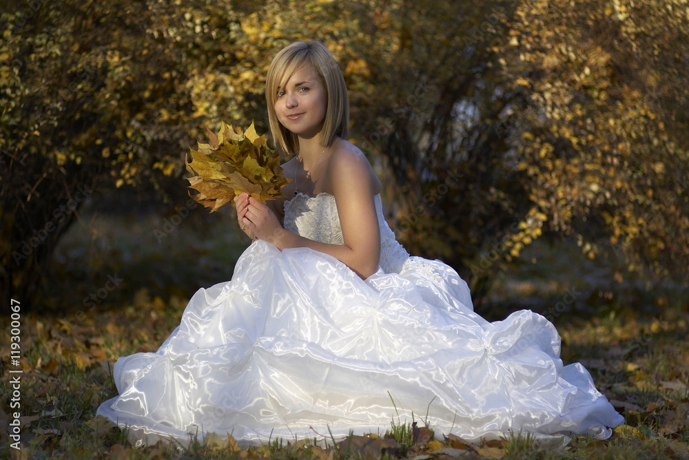 Fototapeta premium Happy Beautiful young bride in white dress sitting in autumn park among fallen leaves/Gorgeous blonde bride in wedding dress with fallen leaves bouquet posing in sunny autumn park