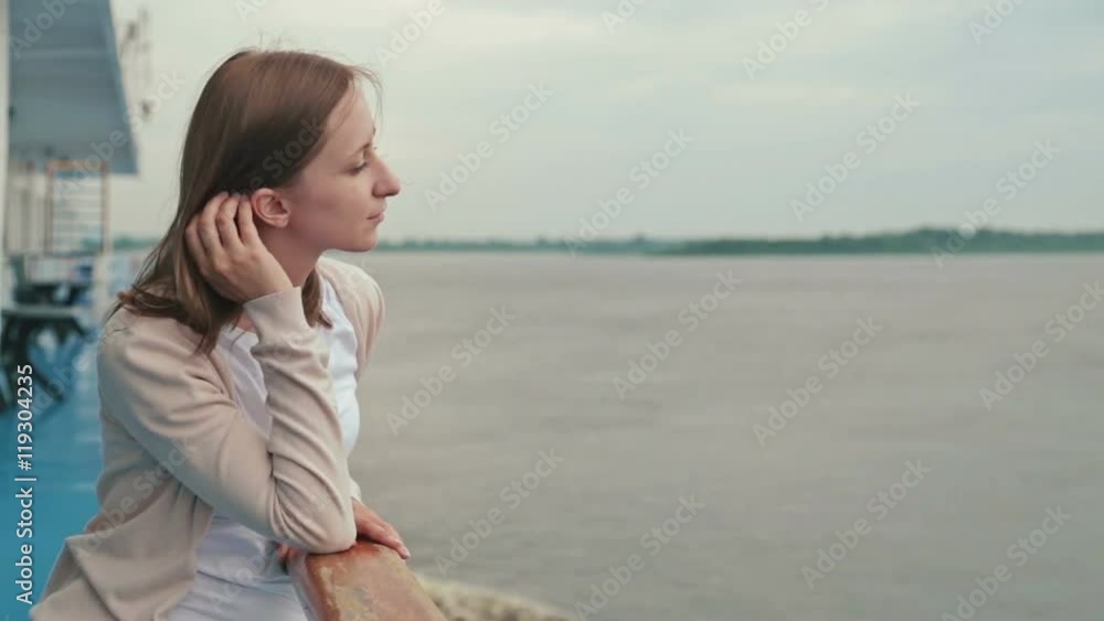 Portrait of young happy beautiful woman on deck of cruise ship. She smiling and looking at river. Rain clouds, overcast. Wind waving her hair. The river can be seen in the background behind her. Portrait of young happy beautiful woman on deck of cruise ship. She smiling and looking at river. Rain clouds, overcast. Wind waving her hair. The river can be seen in the background behind her.