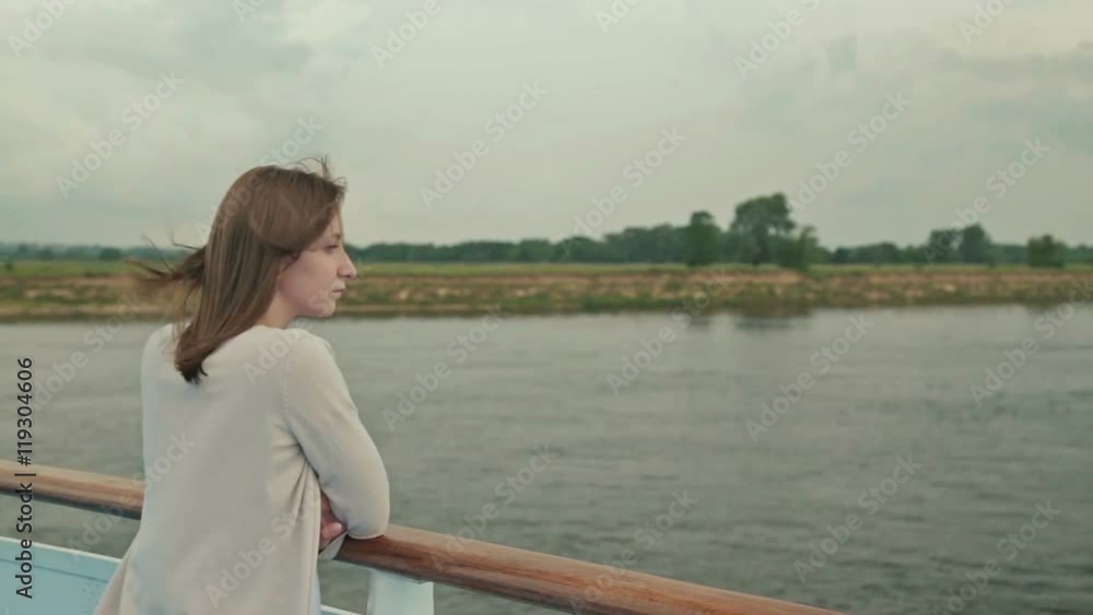 Portrait of young happy beautiful woman on deck of cruise ship. She smiling and looking at river. Rain clouds, overcast. Wind waving her hair. The river can be seen in the background behind her. Portrait of young happy beautiful woman on deck of cruise ship. She smiling and looking at river. Rain clouds, overcast. Wind waving her hair. The river can be seen in the background behind her.