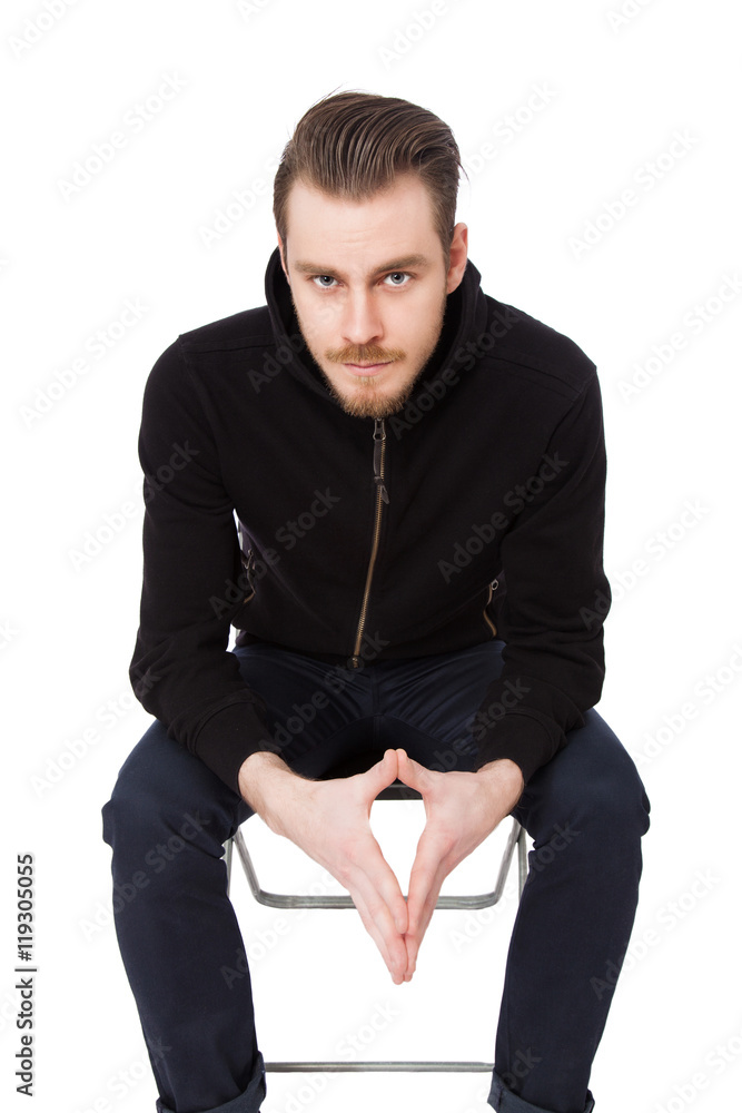 Focused tough man wearing a black hoodie sitting down on a chair staring at camera. White background.