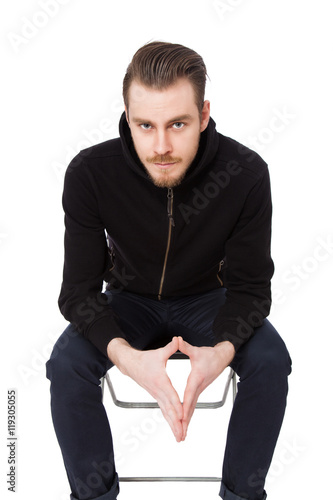 Focused tough man wearing a black hoodie sitting down on a chair staring at camera. White background.