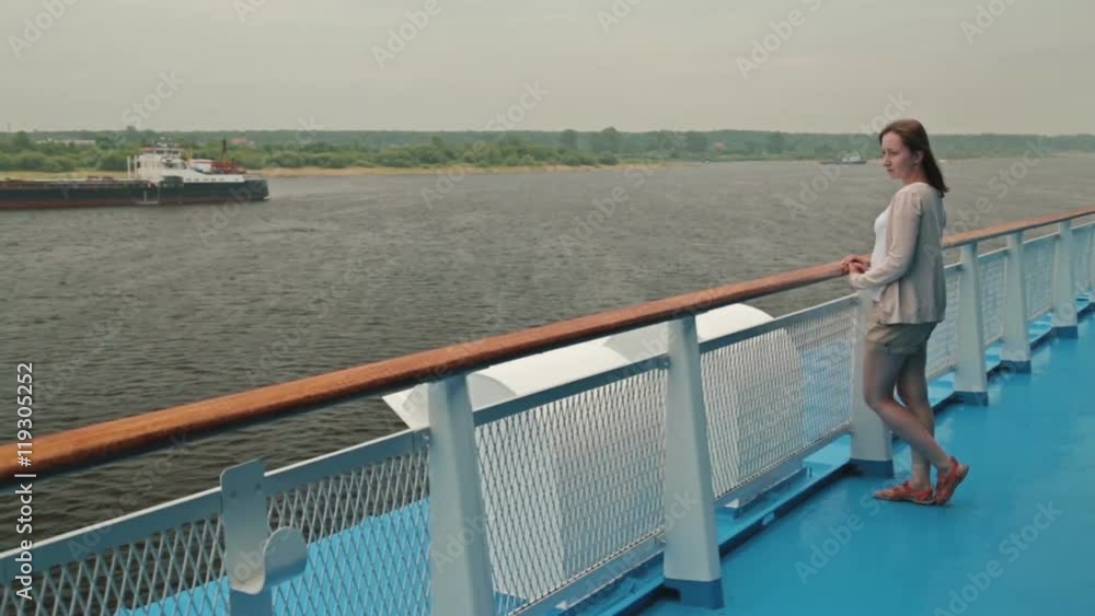 Young happy beautiful woman standing on deck of cruise ship. She smiling and looking at river. Rain clouds, overcast. Wind waving her hair. The riverside can be seen in the background behind her. Young happy beautiful woman standing on deck of cruise ship. She smiling and looking at river. Rain clouds, overcast. Wind waving her hair. The riverside can be seen in the background behind her.