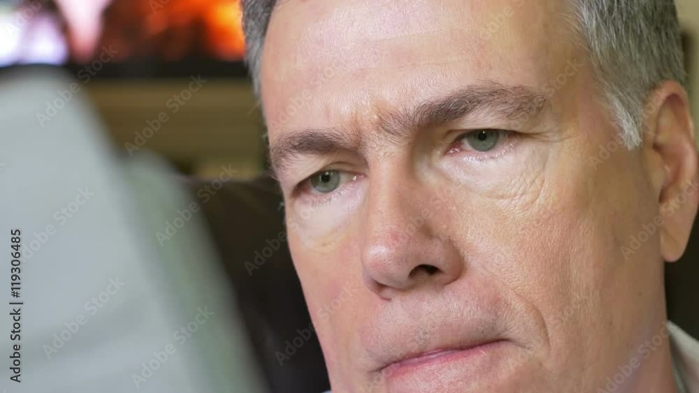 A close-up of a mature Caucasian male manipulating the touch screen of an electronic tablet pc as he reads with TV going in background. A close-up of a mature Caucasian male manipulating the touch screen of an electronic tablet pc as he reads with TV going in background.
