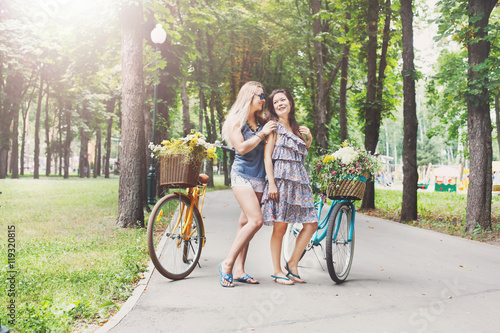 Happy boho chic girls ride together on bicycles in park