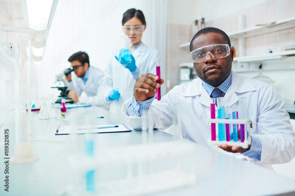 Waist up of male African-American scientist in lab coat and safety goggles  looking at camera holding test tubes, female Asian colleague examining  flask, male Latin-American scientist in background. Stock Photo | Adobe