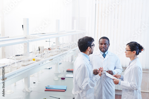 High angle shot of pleased male African-American, male Latin-American and female Asian laboratory scientists in lab coats and glasses discussing scientific research.