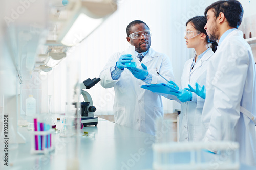 Canvas Print Male African-American laboratory scientist holding flask with blue liquid discussing chemical reaction with male Latin-American and female Asian colleagues