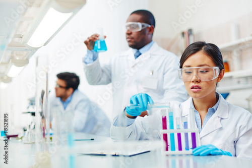 Focused female Asian laboratory scientist in lab coat and safety goggles mixing colored liquid in tubes, male African-American and male Latin-American colleagues working in background.