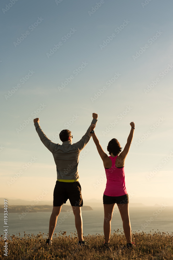 Sporty runner couple rising arms in victory sign after successful training outdoor on the coast facing the sun showing back. Running team success concept. Sporty runner couple rising arms in victory sign after successful training outdoor on the coast facing the sun showing back. Running team success concept.