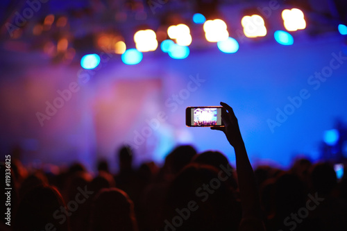 Back lit image of crowd at music concert before stage, focus on one fan holding cellphone high above heads to take picture of memorable moment