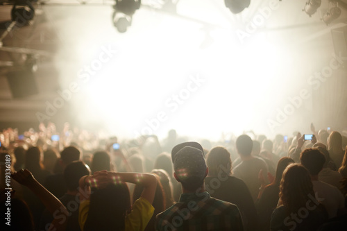 Rear view image of large crowd in front stage in bright light, watching music performance and dancing, focus on young couple in foreground