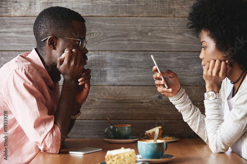 Fotografie Profile of young dark-skinned couple sitting at restaurant: man looking bored an