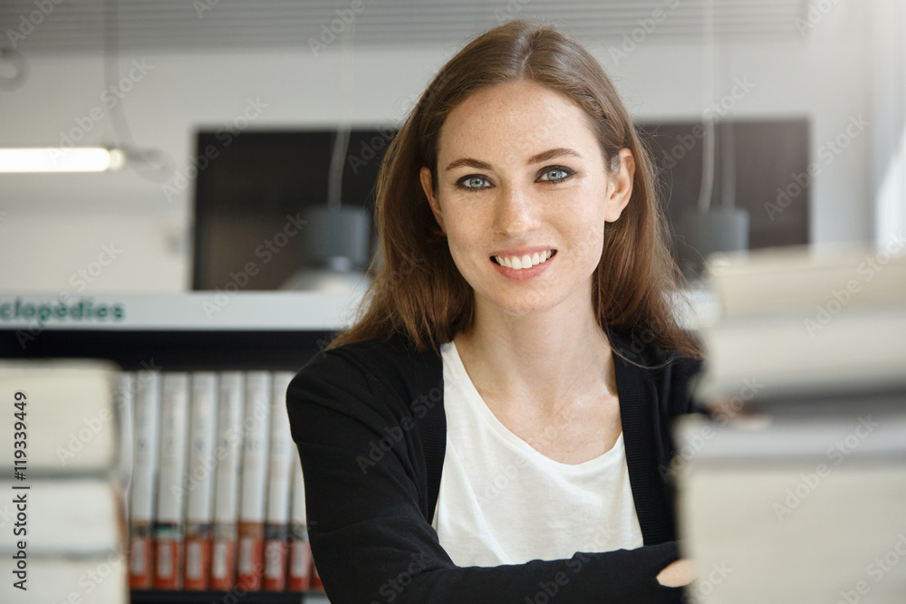 Close up shot of attractive brunette female professor with freckles wearing black jacket, smiling while working on her lecture at university library after classes, surrounded by piles of books